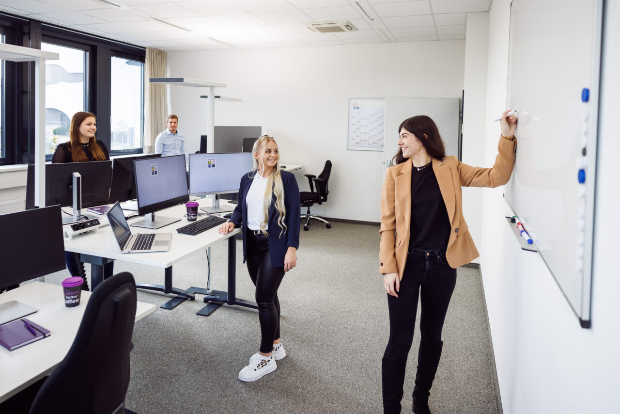 Das Bild zeigt 4 Menschen in einem Büro zusammen. Drei der Menschen stehen vor Ihren Schreibtischen mit Laptop und Monitoren, während eine der vier Personen an einer Tafel steht und etwas aufschreiben möchte.