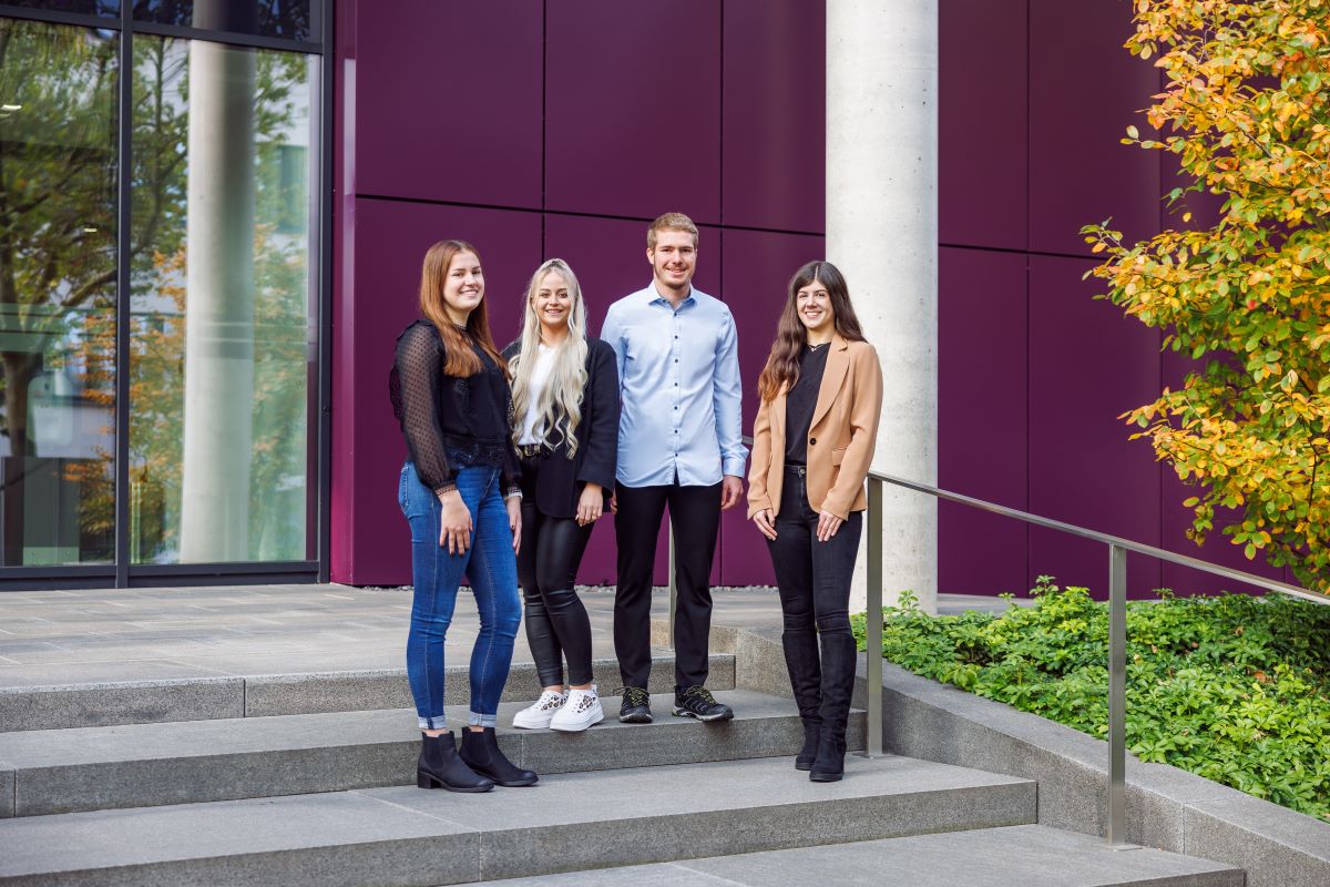 Das Bild zeigt vier junge Erwachsene die auf einer Treppe stehen und in die Kamera lächeln.