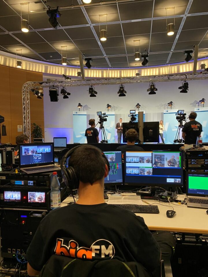 Das Bild zeigt eine bigFM Produktion. Im Hintergrund stehen zwei Moderatoren welche vom bigFM Team gefilmt werden. Im Vordergrund sieht man den Technik Bereich wo Ton, Licht und Bild beobachtet werden.
