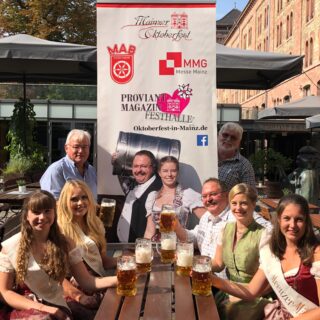 Das Bild zeigt eine Gruppe an Menschen an einem Tisch im Außenbereich sitzen und mit gefüllten Bierkrügen in der Hand. Hinter dem Tisch steht ein Roll-Up des Mainzer Oktoberfestes.