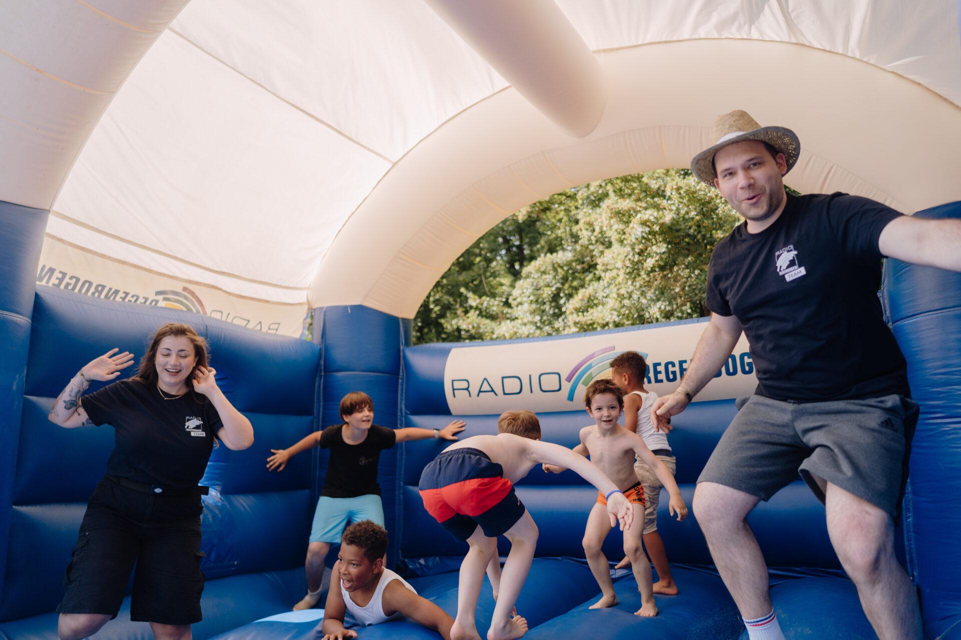Lachende Kinder und die Moderatoren Clara Schardt und Jens Schneider mit Strohhut auf der RADIO REGENBOGEN-Hüpfburg