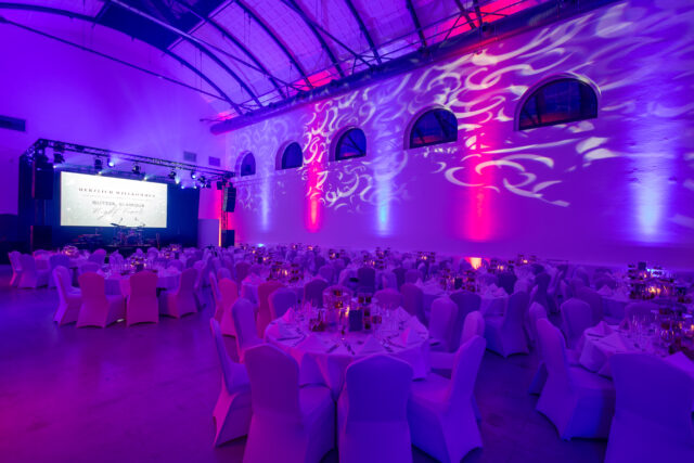 Viele festlich eingedeckte runde Tische mit Hussen über den Stühlen vor einer schön beleuchteten Seitenwand