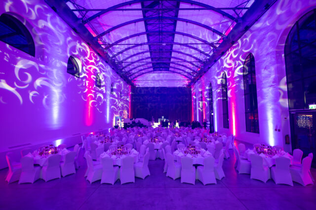 Viele festlich eingedeckte runde Tische mit Hussen über den Stühlen vor einer abgedunkelten Wand in einem gewölbeartigen Raum