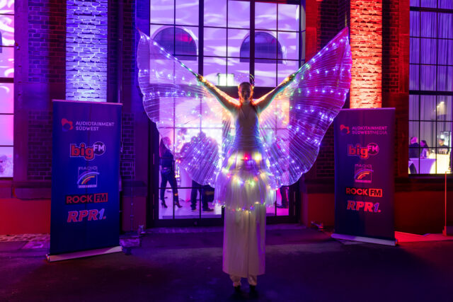 Stelzenläuferin mit weißem Kleid und weißen, beleuchteten Flügeln vor einem Backsteingebäude, die die Arme nach oben nimmt