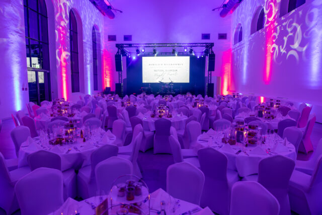 Viele festlich eingedeckte runde Tische mit Hussen über den Stühlen vor einer angestrahlten Leinwand