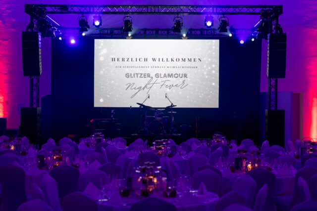 Weiße Leinwand mit Willkommensgruß, davor viele schön eingedeckte Tische mit gediegener Beleuchtung