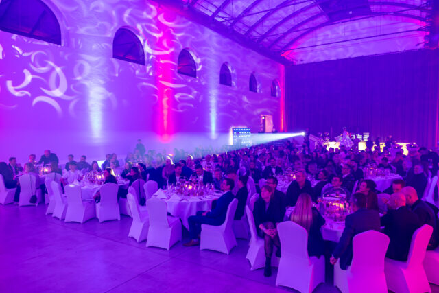 Menschen sitzen in festlicher Kleidung an weiß eingedeckten runden Tischen in einer großen Halle